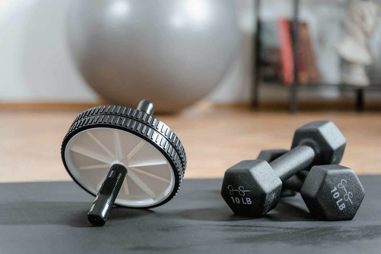 gallery-6 Close-up of ab wheel and dumbbells on a mat in a home gym setting.